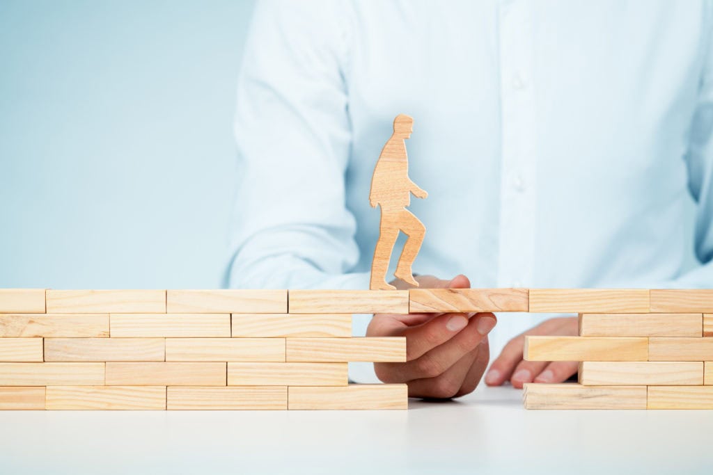 Hand placing a wooden block to bridge a gap for a wooden figure walking across.