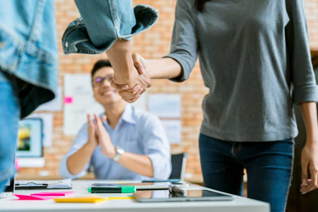 Two colleagues shaking hands over a desk while a coworker applauds in an office.